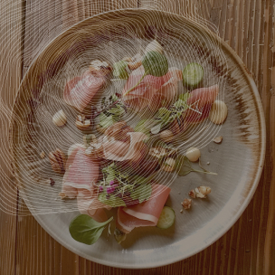 Une assiette en grès contient des tranches de jambon cru, des concombres, des noix, des pousses et des fleurs comestibles. L'assiette est posée sur une table en bois.
