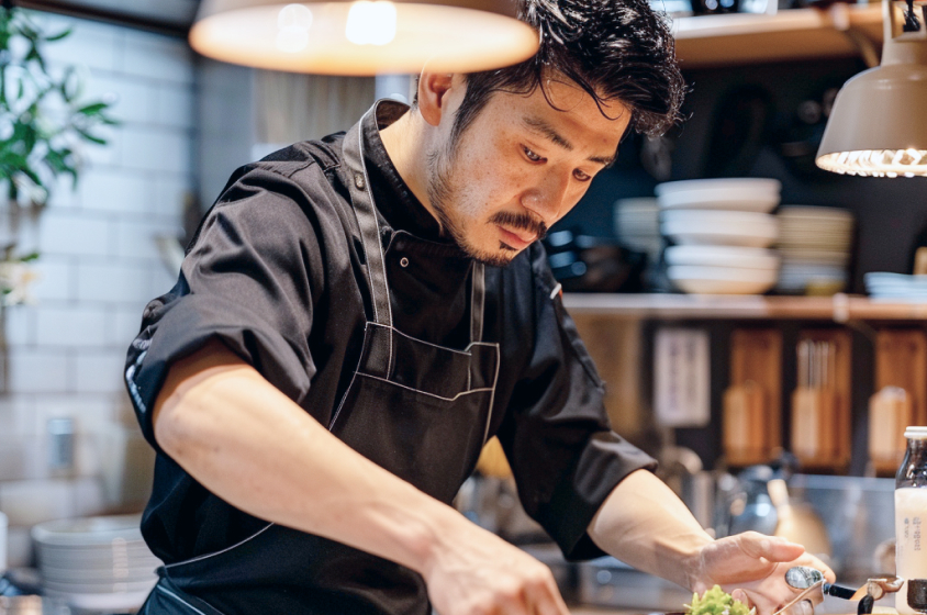 Chef japonais en uniforme noir, intensément concentré sur la préparation d'un plat délicat. Il est debout dans une cuisine bien aménagée avec des ustensiles de cuisine en arrière-plan, éclairé par une lumière chaleureuse au-dessus.