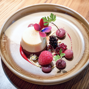 Dessert élégant composé d'un flan crémeux surmonté d'une tranche de poire et décoré de fleurs comestibles, de petites sphères de gelée et de fruits rouges, présenté sur une assiette en céramique rustique.