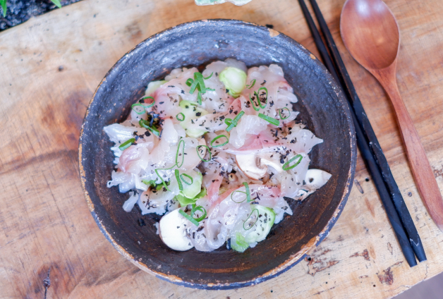 Bol rustique contenant du chirashi, un plat de sashimi varié sur riz vinaigré. Le plat est garni de tranches fines de poisson, de radis, d'oignon vert et de graines de sésame, présenté sur un fond de bois avec des baguettes et une cuillère en bois.