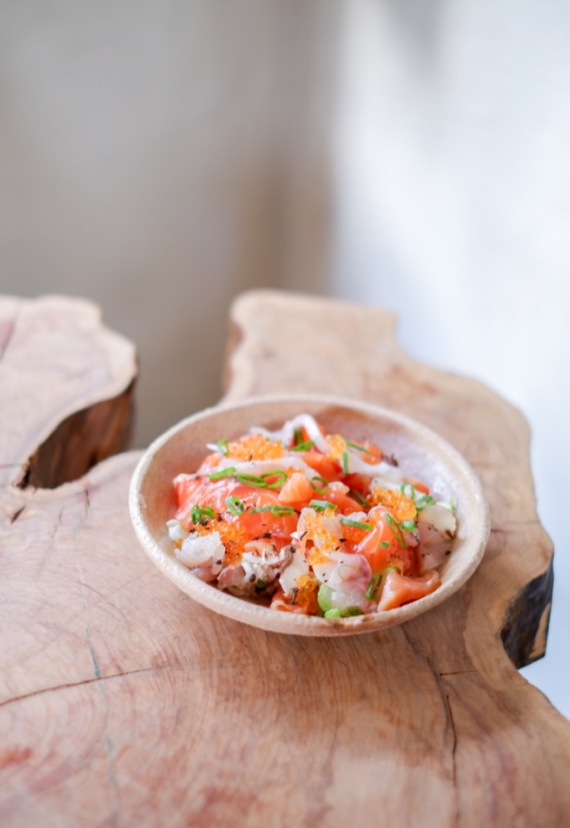 Un bol en argile rustique contenant un mélange de sashimi, incluant du saumon et divers fruits de mer, garni de caviar, d'oignons verts et de fines herbes, présenté sur un support en bois naturel irrégulier."