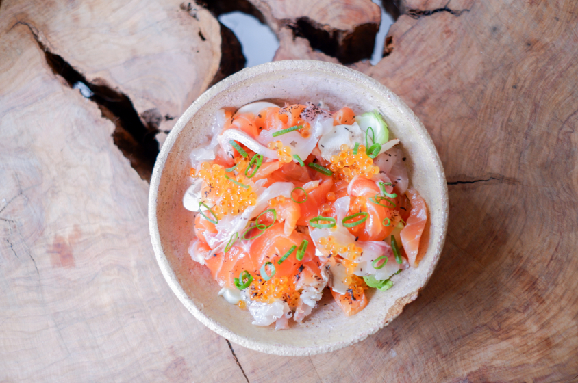 Bol de chirashi vibrant avec sashimi de saumon, œufs de poisson, et fines herbes, garni de crème et servi dans un bol en céramique posé sur une surface en bois naturel, offrant un aperçu de la cuisine japonaise contemporaine.