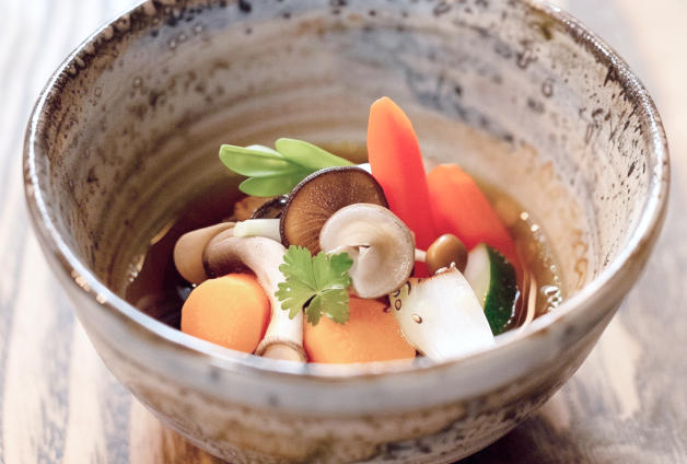 Bol en céramique contenant une soupe claire avec une variété de légumes frais coupés, incluant des carottes, champignons, et des navets, garnie d'herbes fraîches, présentée sur une table en bois pour une touche rustique.