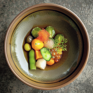 Un bol en céramique marron contient un assortiment de bouchées colorées, dont des légumes verts, des sphères orange et jaunes, une quenelle blanche, des œufs de poisson orange et noirs, une tranche de concombre et un brin d'herbes. Le tout baigne dans un bouillon léger.