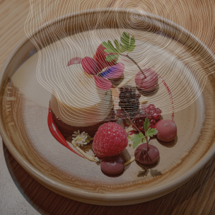 Une assiette en grès marron contient un dessert cylindrique crémeux décoré d'une fleur violette, de framboises, de mûres, de groseilles et de petites fleurs blanches. Des traits de coulis rouge et jaune décorent l'assiette. On devine un motif de feuilles et de cernes d'arbres en filigrane sur l'image.