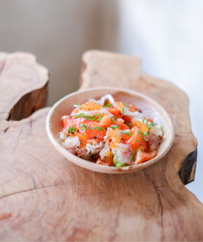 Un bol peu profond en bois contenant des morceaux de saumon cru, des œufs de poisson orange vif et des oignons verts hachés. Le bol repose sur une table en bois.