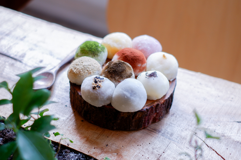 Une tranche de bois servant de plateau à une douzaine de mochis de différentes couleurs et saveurs. Les mochis sont disposés en cercle et saupoudrés de différentes poudres et graines. Une cuillère en métal est posée à côté du plateau. Des feuilles vertes sont visibles au premier plan. Le tout est posé sur une table en bois.