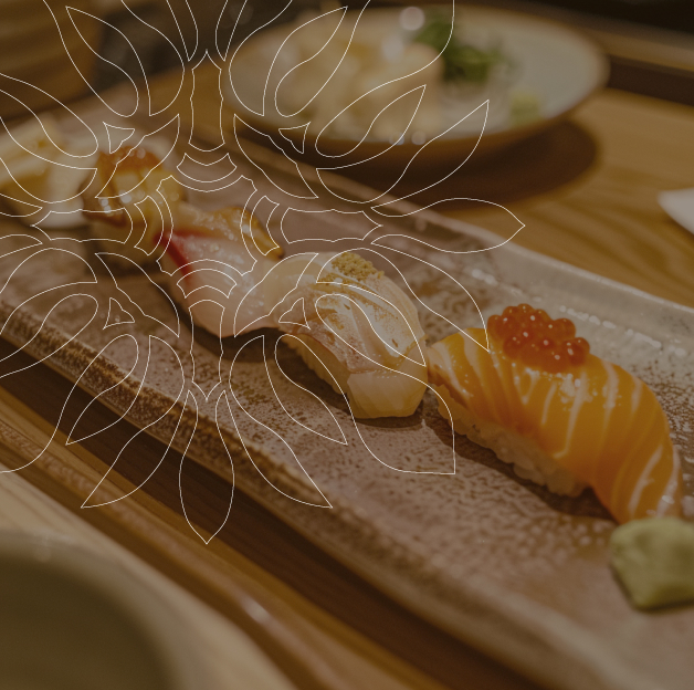 Une assiette rectangulaire en céramique contient trois pièces de sushi différentes. Le sushi le plus à gauche est garni d'un poisson blanc, le sushi du milieu est garni d'un poisson blanc et d'une garniture jaune, et le sushi le plus à droite est garni de saumon et d'œufs de poisson orange. Un wasabi vert pâle est placé à côté du dernier sushi. L'assiette repose sur une table en bois. Une feuille de monstera stylisée est superposée à l'image.