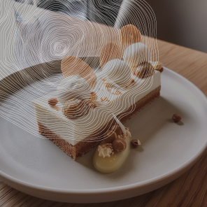 Une part de gâteau rectangulaire est posée sur une assiette blanche. Le gâteau a une base croustillante, une couche de crème et un glaçage blanc. Il est décoré de noisettes, d'amandes et de copeaux de chocolat blanc. Une sauce jaune est disposée à côté du gâteau. L'arrière-plan est flou et présente un motif de lignes courbes blanches.