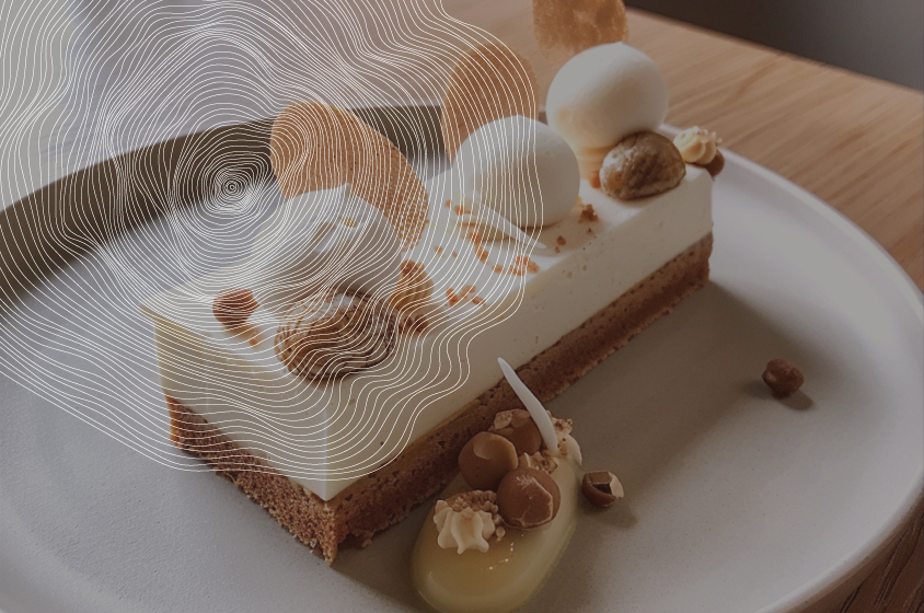 Une part de gâteau rectangulaire est posée sur une assiette blanche. Le gâteau a une base croustillante, une couche de crème et un glaçage blanc. Il est décoré de noisettes, d'amandes et de copeaux de chocolat blanc. Une sauce jaune est disposée à côté du gâteau. L'arrière-plan est flou et présente un motif de lignes courbes blanches.