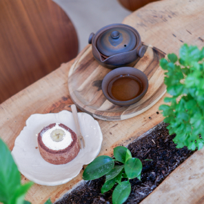 Une table en bois brut avec un dessert, une théière et une tasse de thé posés sur des soucoupes en bois. Il y a une plante verte en pot sur la table. Le dessert est un roulé au chocolat fourré à la crème et à la banane, coupé en deux. La théière et la tasse sont en céramique noire. La table est entourée de plantes vertes.