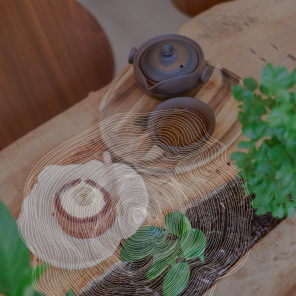 Une table en bois brut avec un dessert, une théière et une tasse de thé posés sur des soucoupes en bois. Il y a une plante verte en pot sur la table. Le dessert est un roulé au chocolat fourré à la crème et à la banane, coupé en deux. La théière et la tasse sont en céramique noire. La table est entourée de plantes vertes.