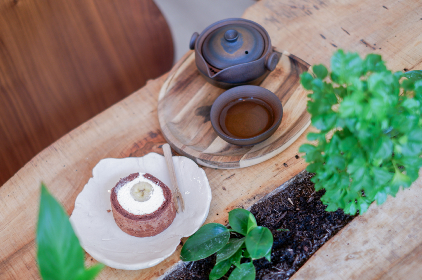 Une table en bois brut avec un dessert, une théière et une tasse de thé posés sur des soucoupes en bois. Il y a une plante verte en pot sur la table. Le dessert est un roulé au chocolat fourré à la crème et à la banane, coupé en deux. La théière et la tasse sont en céramique noire. La table est entourée de plantes vertes.